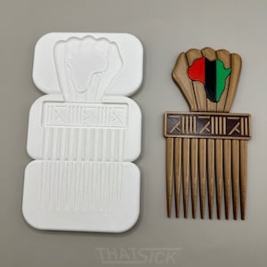 May include: A brown comb with a fist and African continent design, featuring red, black, and green accents. Next to it is a white mold of the comb. The comb has a rectangular handle with vertical teeth.
