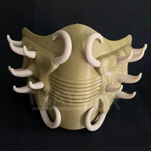 May include: A tan-colored, 3D-printed mask with multiple curved, off-white protrusions. The mask has a textured surface and a series of horizontal ridges across the center. The design is symmetrical and appears to be a decorative or costume piece.