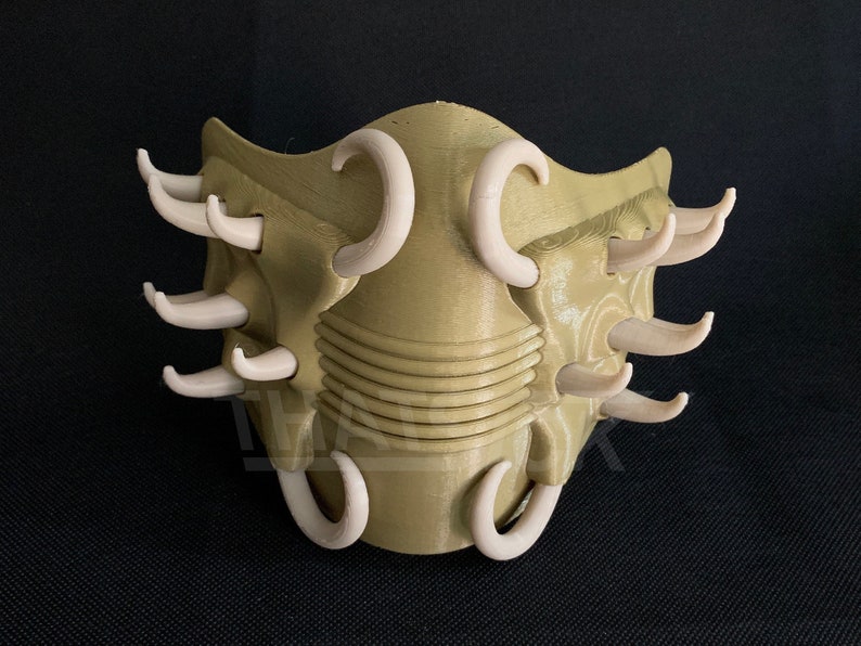 May include: A tan-colored, 3D-printed mask with multiple curved, off-white protrusions. The mask has a textured surface and a series of horizontal ridges across the center. The design is symmetrical and appears to be a decorative or costume piece.