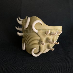 May include: A 3D-printed mask in a tan color, featuring numerous curved, off-white protrusions. The mask has a textured surface and a design that suggests a creature or alien aesthetic. The mask is set against a dark background.