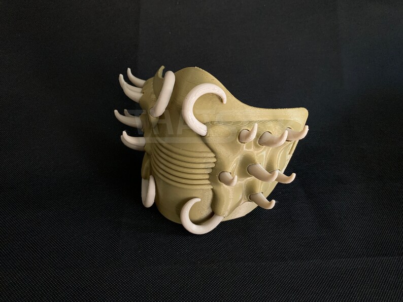May include: A 3D-printed mask in a tan color, featuring numerous curved, off-white protrusions. The mask has a textured surface and a design that suggests a creature or alien aesthetic. The mask is set against a dark background.