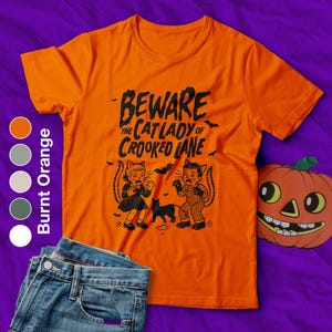 Op de afbeelding: Oranje shirt met de tekst "BEWARE THE CAT LADY OF CROOKED LANE" en een afbeelding van twee katachtige figuren en een zwarte kat. Een decoratieve pompoen en een spijkerbroek staan ook op de afbeelding.