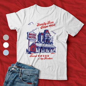 May include: White t-shirt with a graphic of the Bates Motel, text reads "Family Run Since 1960" and "Rated...by Mother." The design is in red, white, and blue, with a vintage, illustrated style. Blue jeans are visible in the foreground.