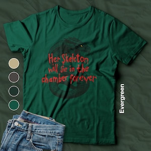 Puede incluir: Camiseta verde esmeralda con cuello redondo y mangas cortas. La camiseta presenta un gráfico de dragón negro con texto rojo que dice "Her Skeleton will lie in the chamber forever". Un par de vaqueros azules es visible en la imagen.