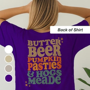 May include: A purple t-shirt with the text "BUTTER BEER PUMPKIN PASTIES & HOGS MEADE" in brown, orange, and green lettering. The shirt has a retro design and is a fun, casual wear item.