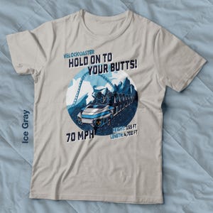Puede incluir: Camiseta gris hielo con un gráfico de montaña rusa y el texto "VELOCKOASTER HOLD ON TO YOUR BUTTS!". El diseño incluye la velocidad de la montaña rusa a 112.65 km/h, la altura a 47.24 m y la longitud a 1432.56 m.