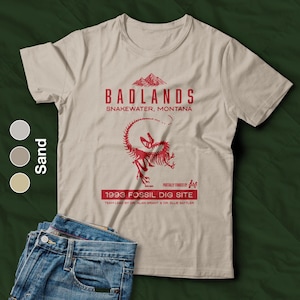 May include: Sand-colored t-shirt with a red graphic of a dinosaur skeleton and text that reads "BADLANDS SNAKEWATER, MONTANA 1993 FOSSIL DIG SITE". The shirt is paired with blue jeans.