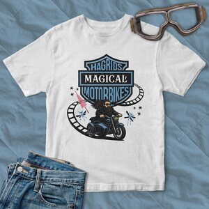 May include: White t-shirt featuring a graphic of a bearded man riding a blue motorcycle with the text "Hagrids Magical Motorbikes". The design includes a pink umbrella and stylized insects. Goggles are visible in the upper right corner.