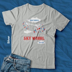 Op de afbeelding: Zilverkleurig t-shirt met een afbeelding van de Sky Whirl-attractie van Great America. Het ontwerp bevat de tekst "SKY WHIRL" en "1976-2000" in rood en wit. De woorden "Great America" staan boven de attractie in een wolk.