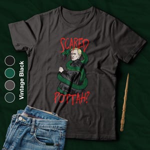 Op de afbeelding: Vintage zwart t-shirt met een afbeelding van een personage met een groene slang en de rode tekst "Scared Pottah?". Een houten toverstaf en een spijkerbroek zijn ook zichtbaar.