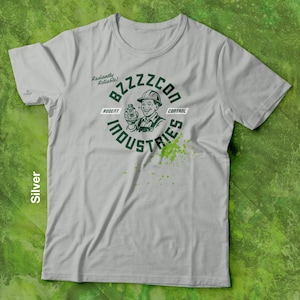 Op de afbeelding: Zilverkleurig T-shirt met een groene afbeelding van een lachende man met een helm die een fles vasthoudt, met de tekst "BZZZZCON INDUSTRIES" en "Rodent Control". De woorden "Radiantly Reliable!" staan erboven.