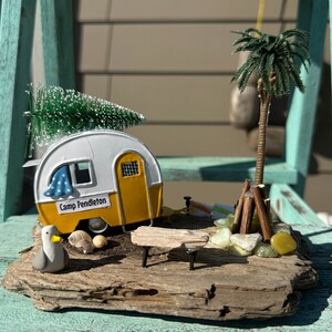 Pode incluir: Um autocaravana em miniatura amarelo e branco com o texto "Camp Pendleton" no lado, uma pequena palmeira e uma fogueira em miniatura numa base de madeira à deriva.