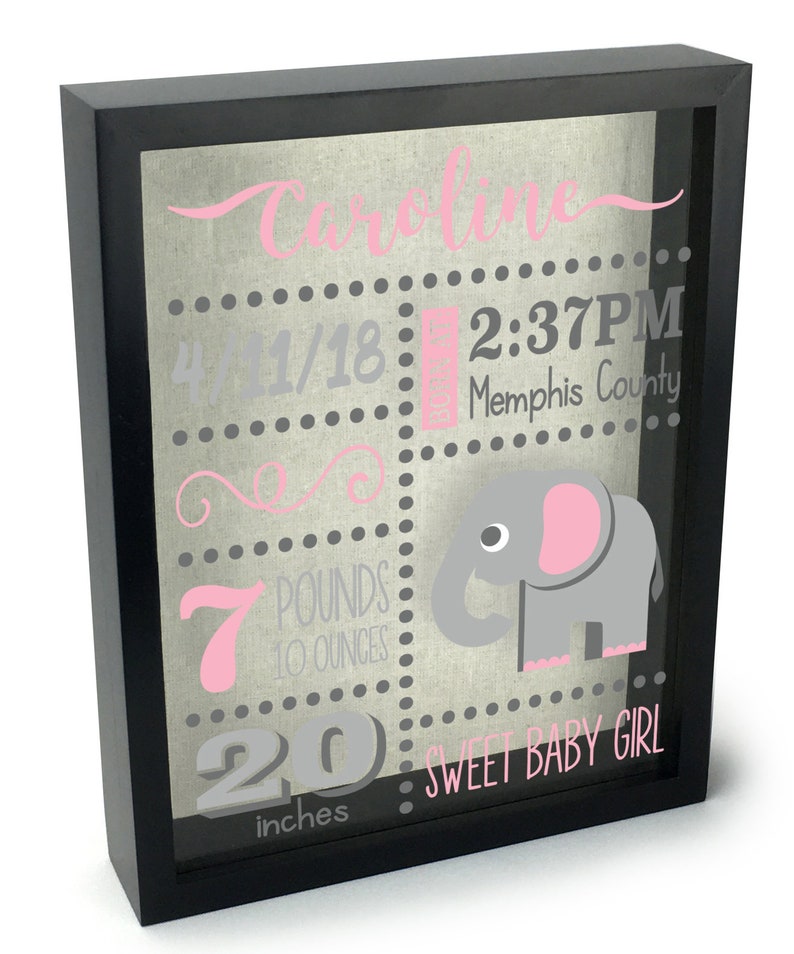 May include: A black framed shadow box with a grey and pink elephant design. The box includes the name "Caroline", the date "4/11/18", the time "2:37 PM", the location "Memphis County", the weight "7 pounds 10 ounces", and the length "20 inches". The text "Sweet Baby Girl" is also included.