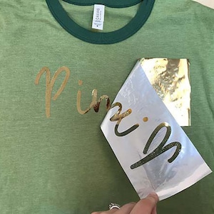 May include: A green t-shirt with a gold foil design that says "Pink" on it. The gold foil is being peeled off of a clear sheet.