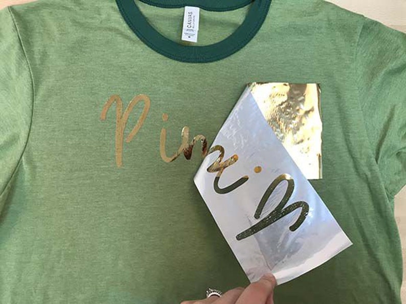 May include: A green t-shirt with a gold foil design that says "Pink" on it. The gold foil is being peeled off of a clear sheet.