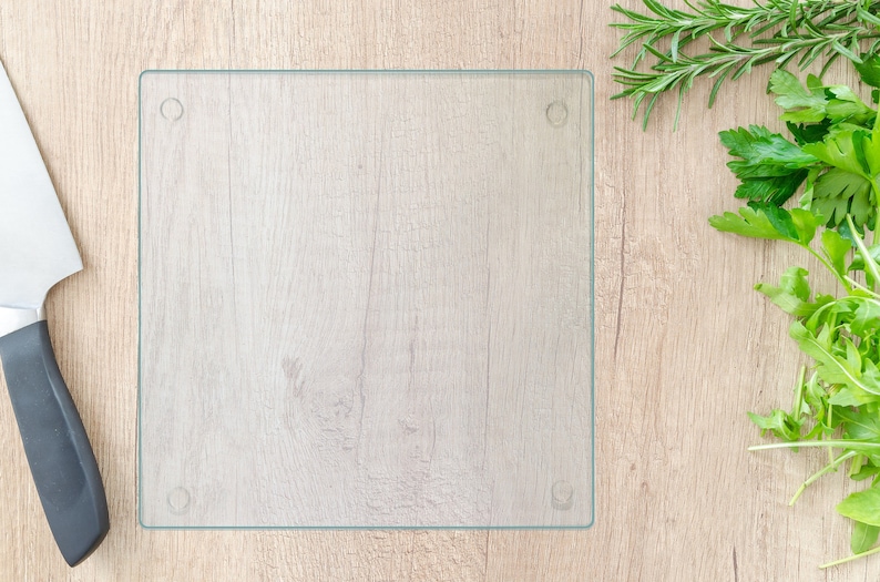 May include: A clear glass cutting board on a light wood surface. There is a black chef's knife and a bunch of fresh herbs to the right of the board.