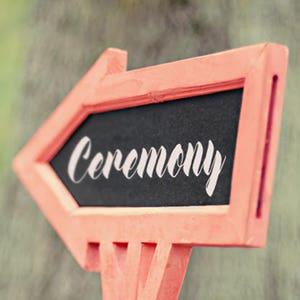 Op de afbeelding: Een rood houten pijlschild met een zwarte krijtbordinzet waarop in witte cursieve letters "Ceremonie" staat.