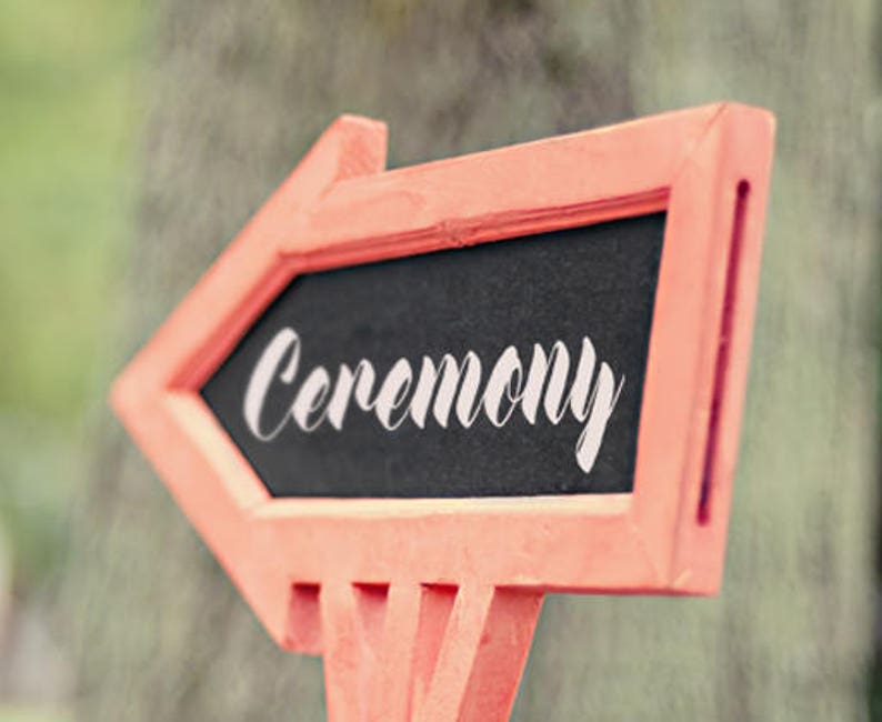 Op de afbeelding: Een rood houten pijlschild met een zwarte krijtbordinzet waarop in witte cursieve letters "Ceremonie" staat.