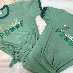 May include: Two green t-shirts with a gold glitter design that says "Pinch Proof" with green shamrocks. The shirts have a green ringer.