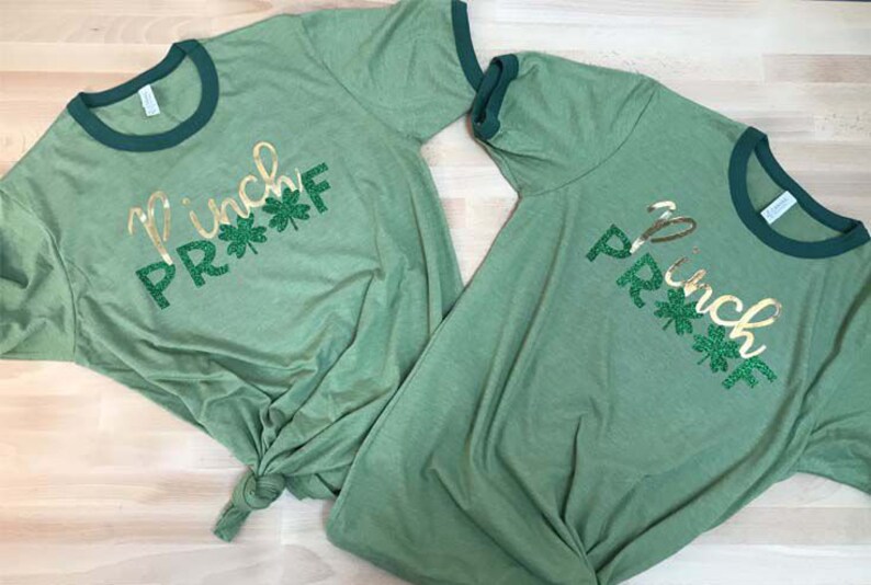 May include: Two green t-shirts with a gold glitter design that says "Pinch Proof" with green shamrocks. The shirts have a green ringer.