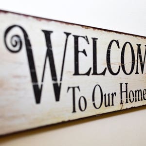 May include: A rectangular wooden sign with the words "WELCOME To Our Home" in black lettering. The sign has a distressed, vintage look with a white background and brown edges. Decorative swirls accent the word "WELCOME".