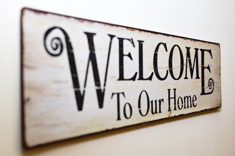 May include: A rectangular wooden sign with the words "WELCOME To Our Home" in black lettering. The sign has a distressed, vintage look with a white background and brown edges. Decorative swirls accent the word "WELCOME".