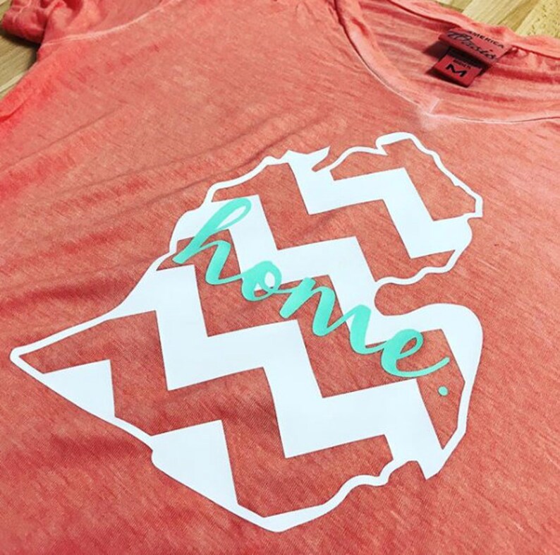 May include: A coral colored t-shirt with a white outline of the state of Michigan with a chevron pattern inside. The word "home" is written in teal inside the state outline.