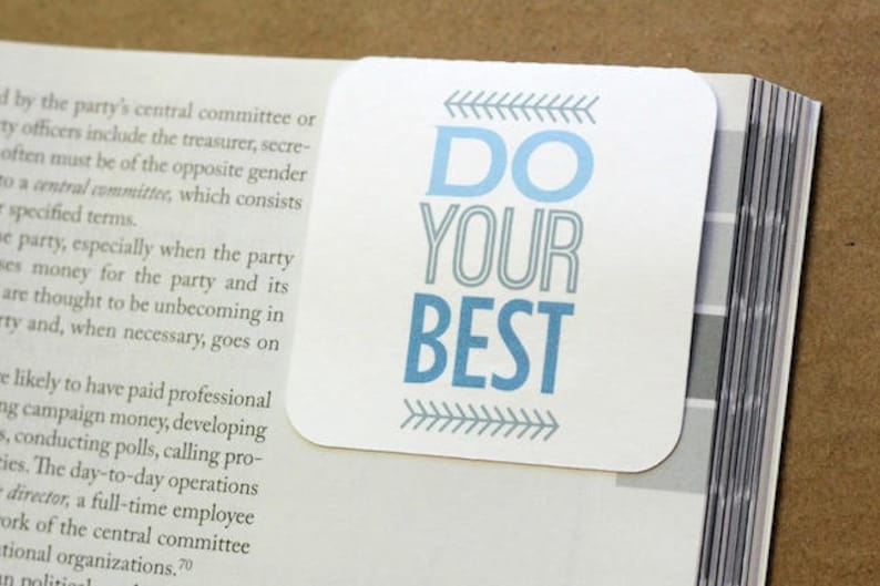 以下が含まれることがあります： 「DO YOUR BEST」という青い文字が書かれた白いしおり。文字はグレーで縁取られており、文字の両側にグレーの矢印があります。