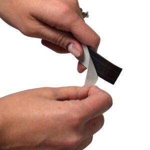 Pu&ograve; includere: Una mano sta rimuovendo il rivestimento protettivo da un rotolo di nastro magnetico nero.