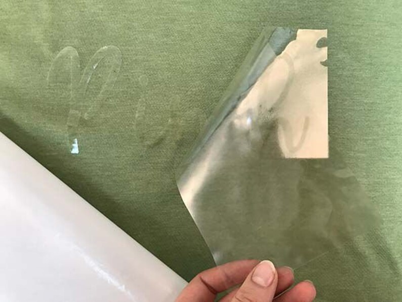 May include: A clear adhesive sheet is being peeled off of a green fabric surface. The adhesive sheet has a design on it that is visible through the sheet.