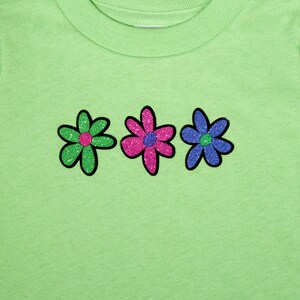 May include: A light green t-shirt with three glitter flowers. The flowers are green, pink, and blue.