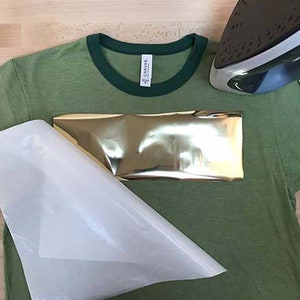 May include: A green t-shirt with a gold metallic heat transfer design. A white heat press sheet is placed over the design and an iron is positioned above the sheet.