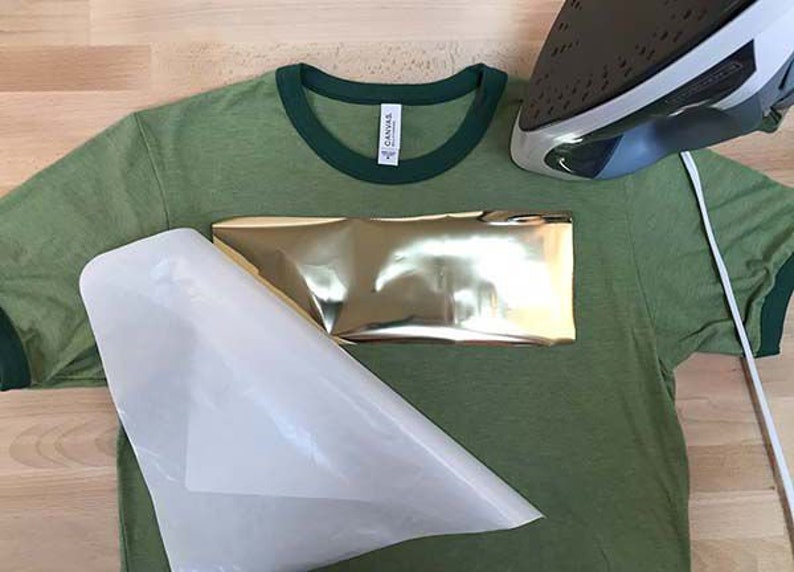 May include: A green t-shirt with a gold metallic heat transfer design. A white heat press sheet is placed over the design and an iron is positioned above the sheet.