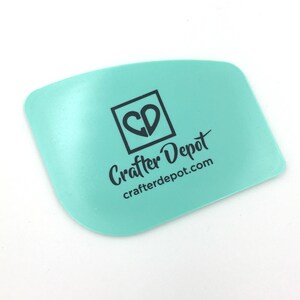May include: A light blue plastic scraper with a heart-shaped logo that says "CD" inside a square. The text "Crafter Depot" and "crafterdepot.com" is printed below the logo.