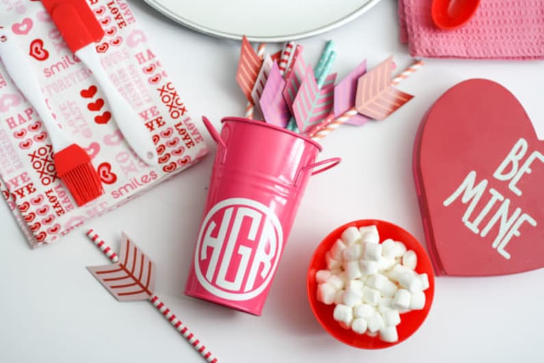 Op de afbeelding: Een roze metalen emmer met een wit monogram "HGR" aan de voorkant. De emmer is omringd door Valentijnsdag-artikelen, waaronder rood-wit gestreepte rietjes met papieren pijlen, een rood hartvormig bord met de tekst "Be Mine" en een kom met witte marshmallows.