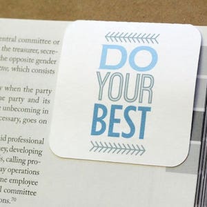 Pu&ograve; includere: Un segnalibro bianco con testo blu che recita "DO YOUR BEST". Il testo &egrave; contornato di grigio e ci sono frecce grigie su entrambi i lati del testo.