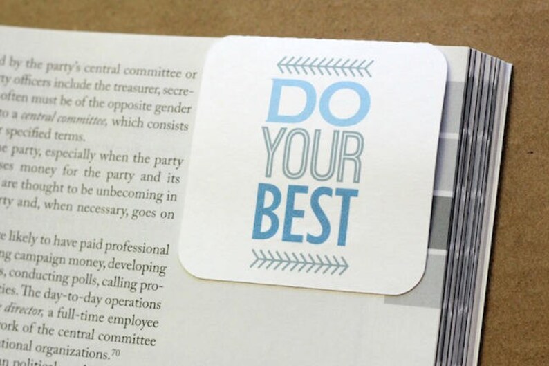 Pu&ograve; includere: Un segnalibro bianco con testo blu che recita "DO YOUR BEST". Il testo &egrave; contornato di grigio e ci sono frecce grigie su entrambi i lati del testo.