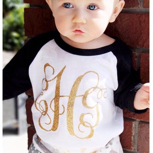 May include: A baby wearing a white and black baseball tee with a gold glitter letter 'H' on the front. The baby is also wearing a white and gray patterned skirt and a white headband with gold polka dots.