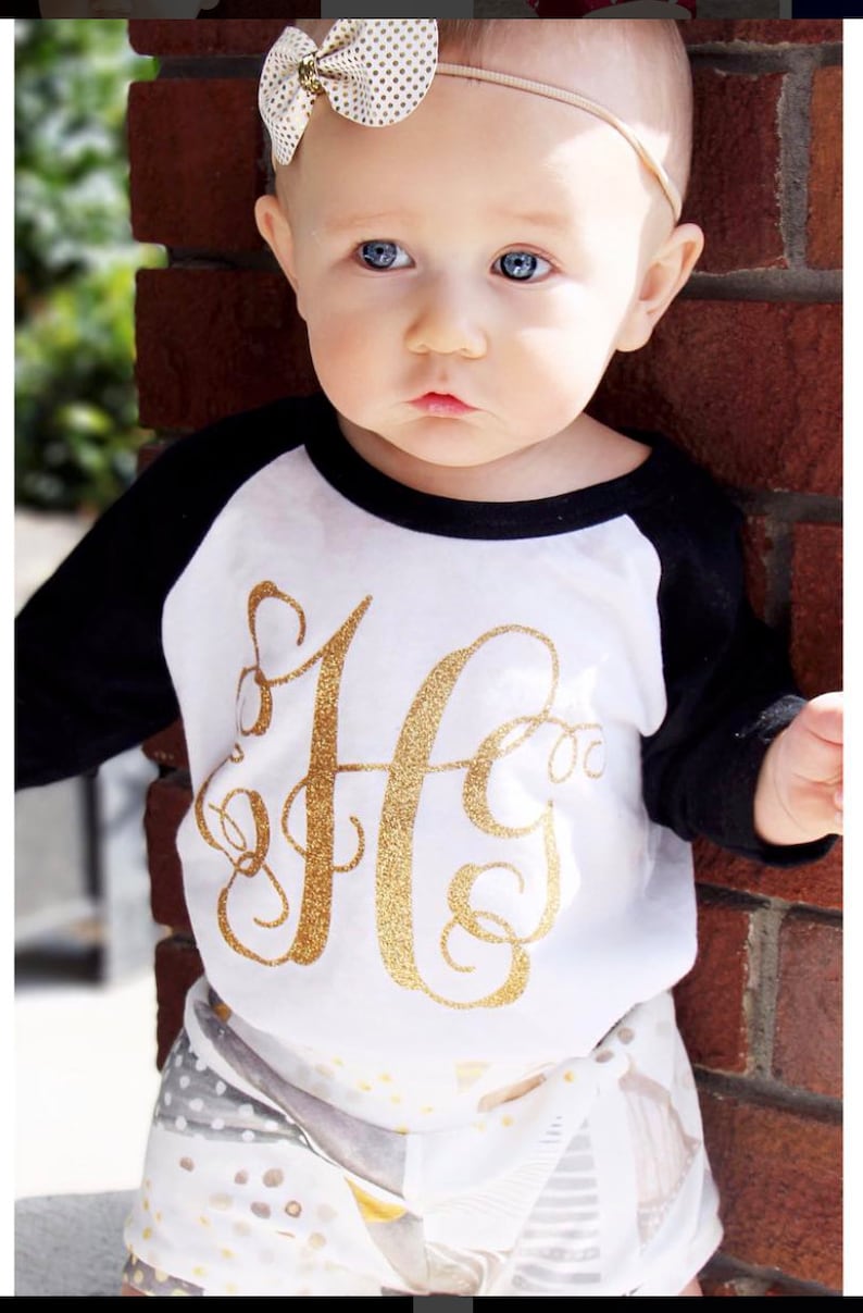 May include: A baby wearing a white and black baseball tee with a gold glitter letter 'H' on the front. The baby is also wearing a white and gray patterned skirt and a white headband with gold polka dots.