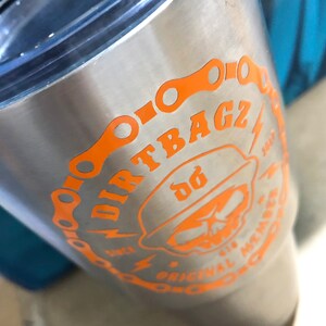 Op de afbeelding: Een roestvrijstalen tumbler met een oranje afbeelding van een schedel die een hoed draagt met de tekst "DIRTBAGZ" en "ORIGINAL MEMBER" rond de schedel. De afbeelding bevat ook een kettingschakelontwerp.