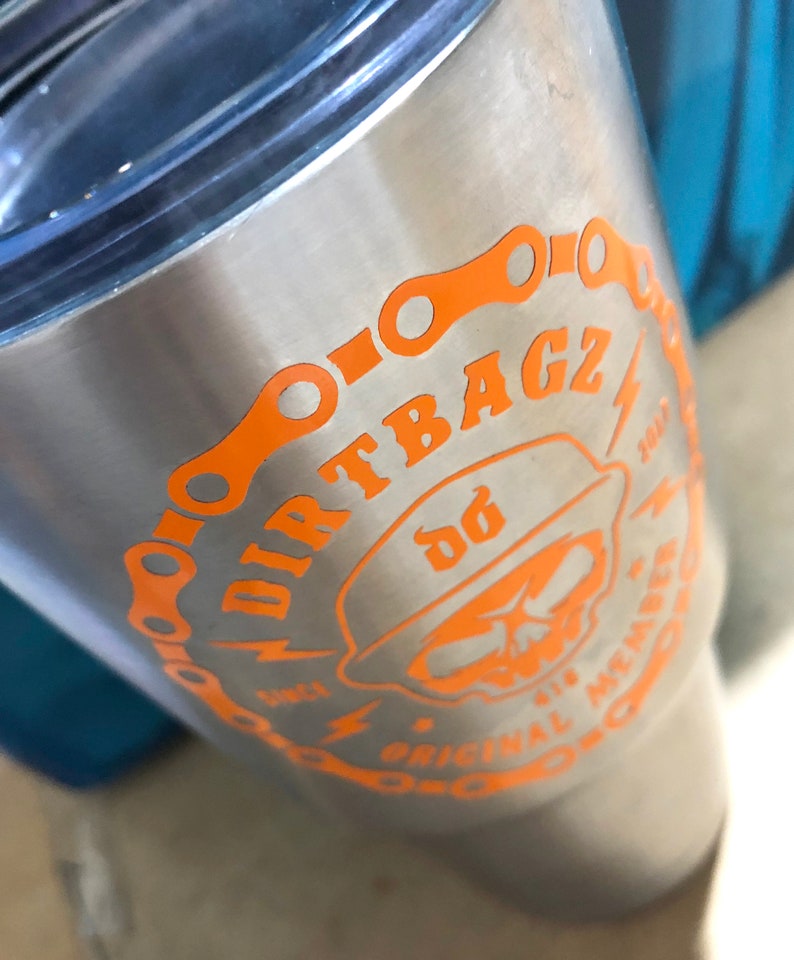 Op de afbeelding: Een roestvrijstalen tumbler met een oranje afbeelding van een schedel die een hoed draagt met de tekst "DIRTBAGZ" en "ORIGINAL MEMBER" rond de schedel. De afbeelding bevat ook een kettingschakelontwerp.
