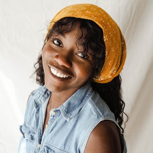 May include: A woman with curly dark hair is wearing a yellow bandana with a white pattern and a denim sleeveless shirt. She is smiling.