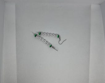 May include: A pair of handmade earrings spelling out the word "SCHOOL" with white and green letter beads. The earrings have silver-colored fish hook ear wires and are displayed on a white background.