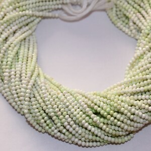 May include: A collection of pale green, small, round beads, likely gemstones or pearls, are arranged in multiple strands. The beads have a subtle sheen and are tightly strung together, creating a textured, organic appearance. The strands are gathered at the top.