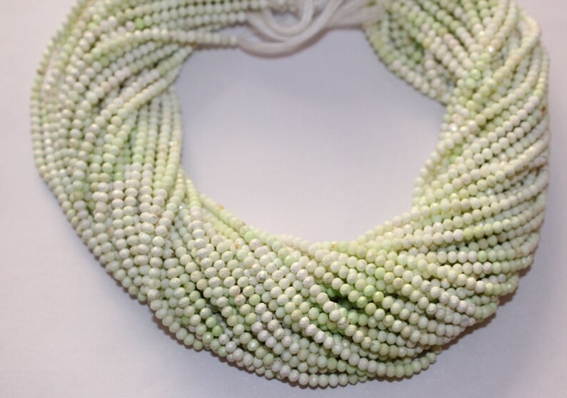 May include: A collection of pale green, small, round beads, likely gemstones or pearls, are arranged in multiple strands. The beads have a subtle sheen and are tightly strung together, creating a textured, organic appearance. The strands are gathered at the top.