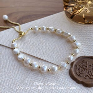 May include: A delicate pearl bracelet with a gold chain and accents. The bracelet features irregularly shaped white pearls interspersed with small gold beads. The adjustable clasp is also gold. The text "DiversityNotions" and "Who says my dream has to stay dream?" is visible.