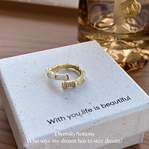 May include: A gold-colored ring with white enamel accents and small clear stones. The ring is on a white box with the text "With you, life is beautiful" and "Who says my dream has to stay dream?"
