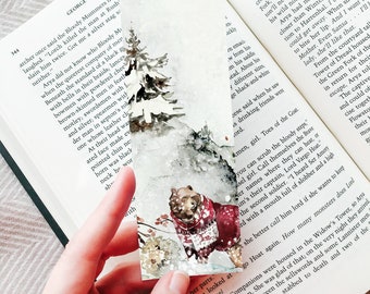 May include: A rectangular paper bookmark featuring a watercolor illustration of a brown bear wearing a red and white patterned sweater. The scene includes a dark green pine tree, snowy ground, and small floral accents against a light background.