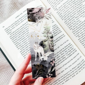 May include: A rectangular bookmark featuring a watercolor illustration of a grey wolf standing in a forest with pine trees, dark abstract shapes, gold geometric lines, and small gold splatters against a white and grey background.