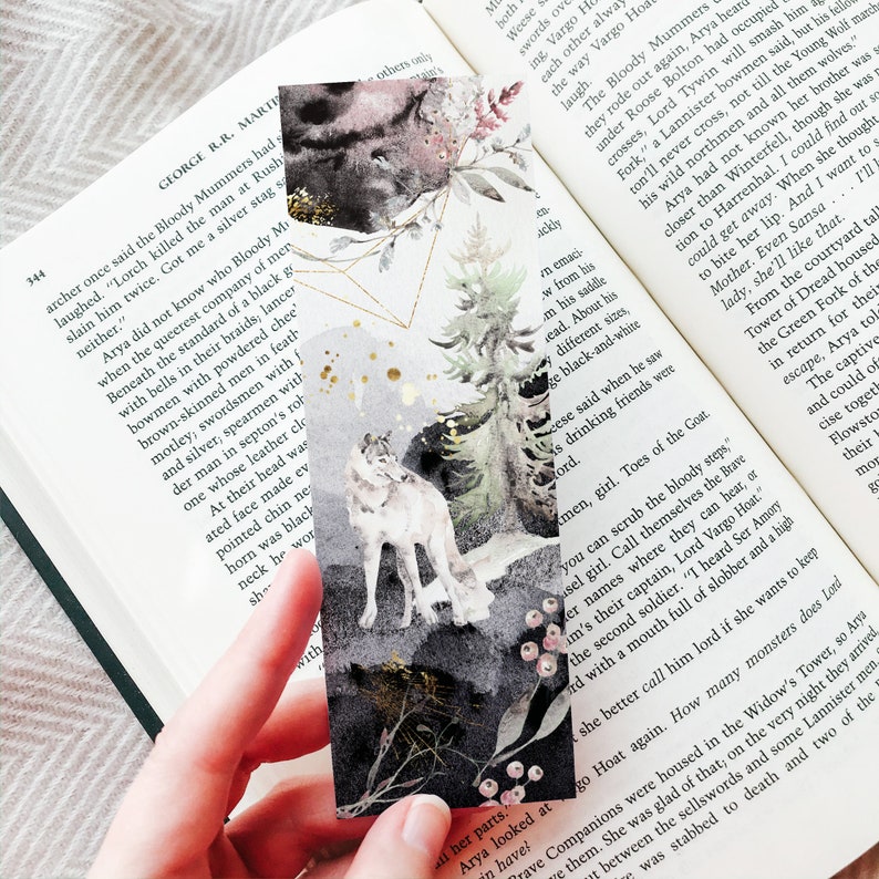 May include: A rectangular bookmark featuring a watercolor illustration of a grey wolf standing in a forest with pine trees, dark abstract shapes, gold geometric lines, and small gold splatters against a white and grey background.